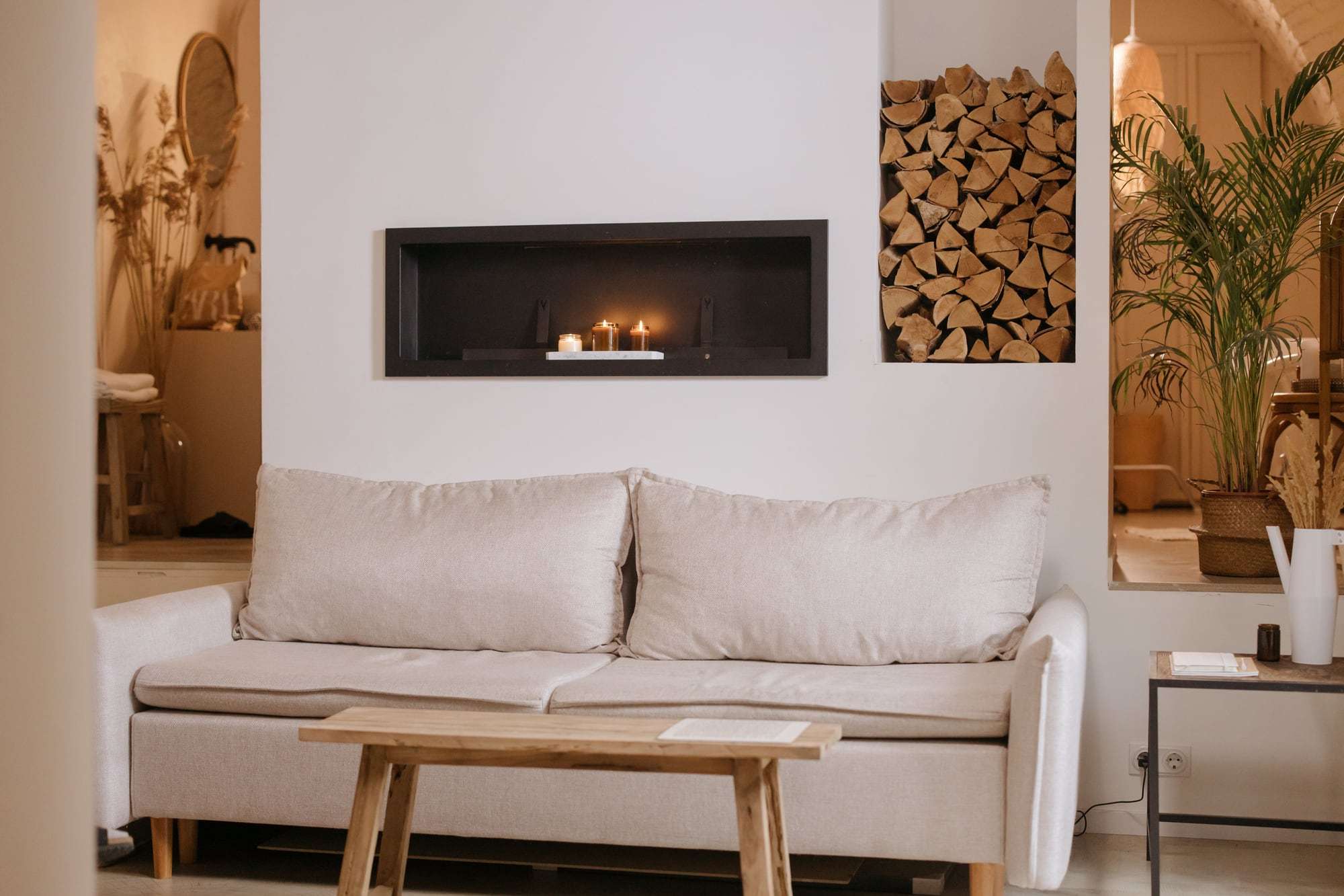 Helles Wohnzimmer mit weißem Sofa, Holztisch und Kaminnische mit gestapeltem Holz